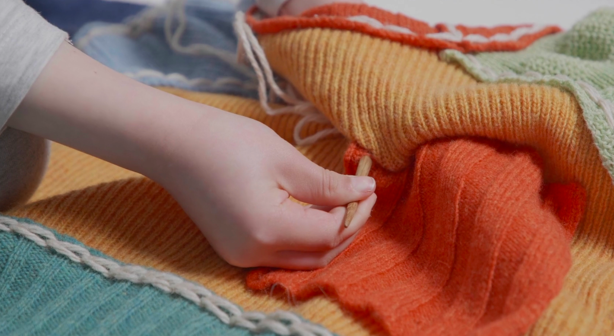 A child's hand using a wooden needle to stitch panels of wool