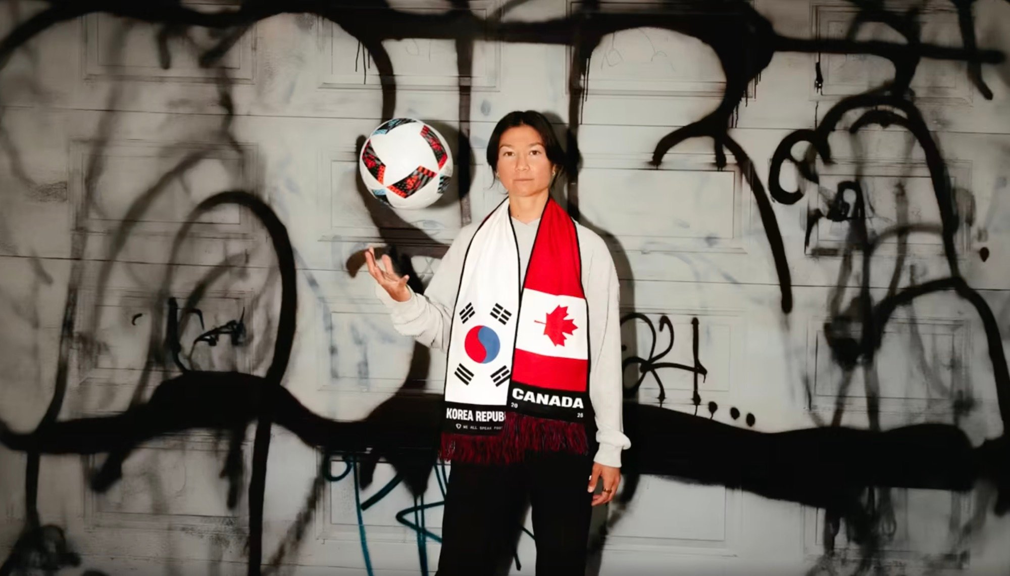 A person wearing a dual-sided soccer scarf with the South Korean flag on one side and the Canadian flag on the other, tossing a ball in the air against a graffiti-covered wall
