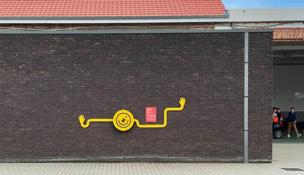 Yellow HAPPY Speeltijd wheel with long, extended arms on a brick school wall