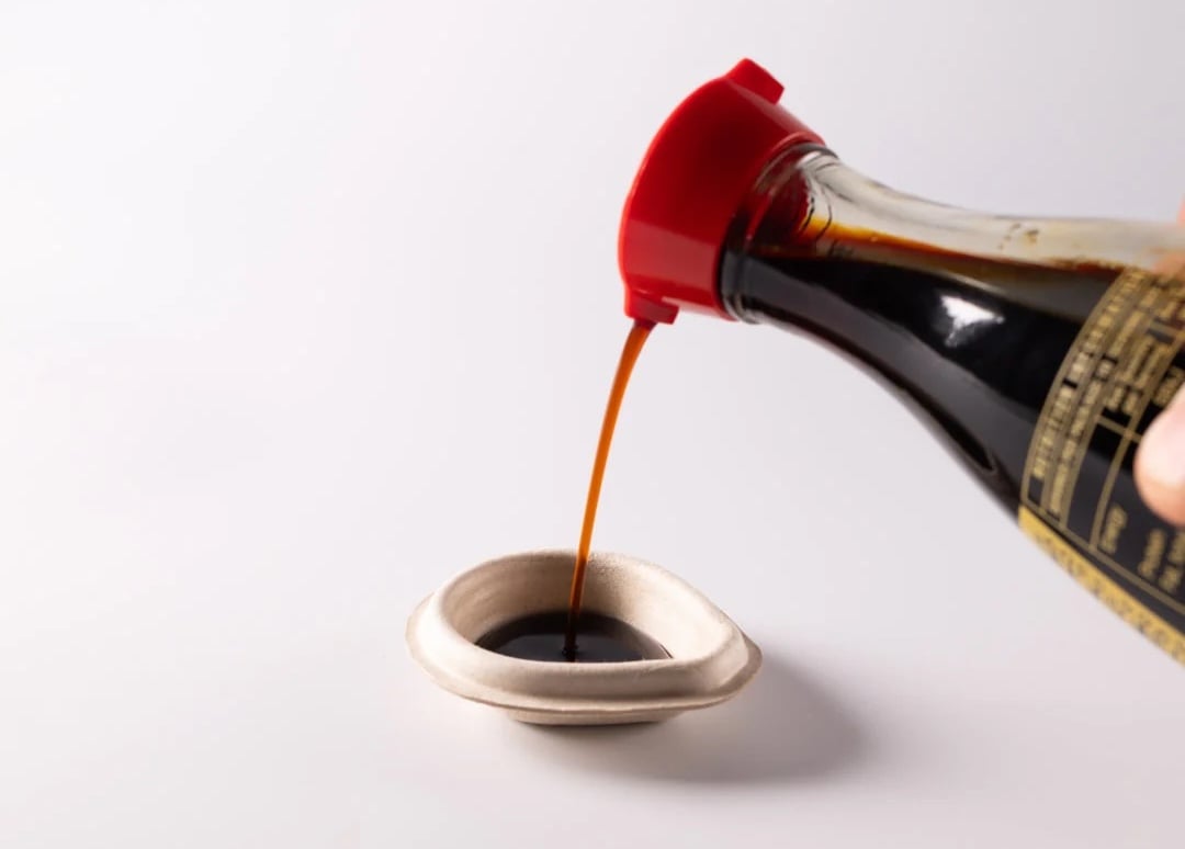 Soy sauce being poured from a glass bottle into a beige, fish-shaped Holy Carp! compostable container made from bagasse plant pulp