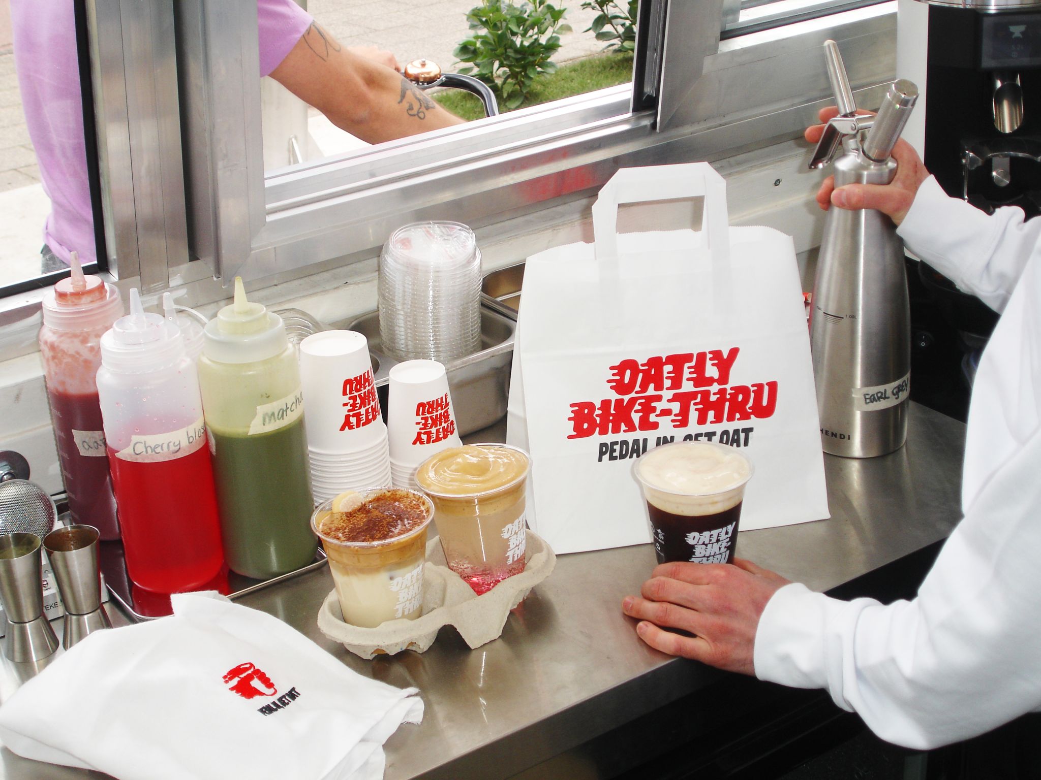 Behind the Oatly Bike-Thru counter, a barista hands a cyclist a cold oat drink topped with foam while three signature drinks sit ready in a cup carrier, alongside labeled syrup bottles for cherry blossom and matcha, branded cups, and an Oatly Bike-Thru takeaway bag