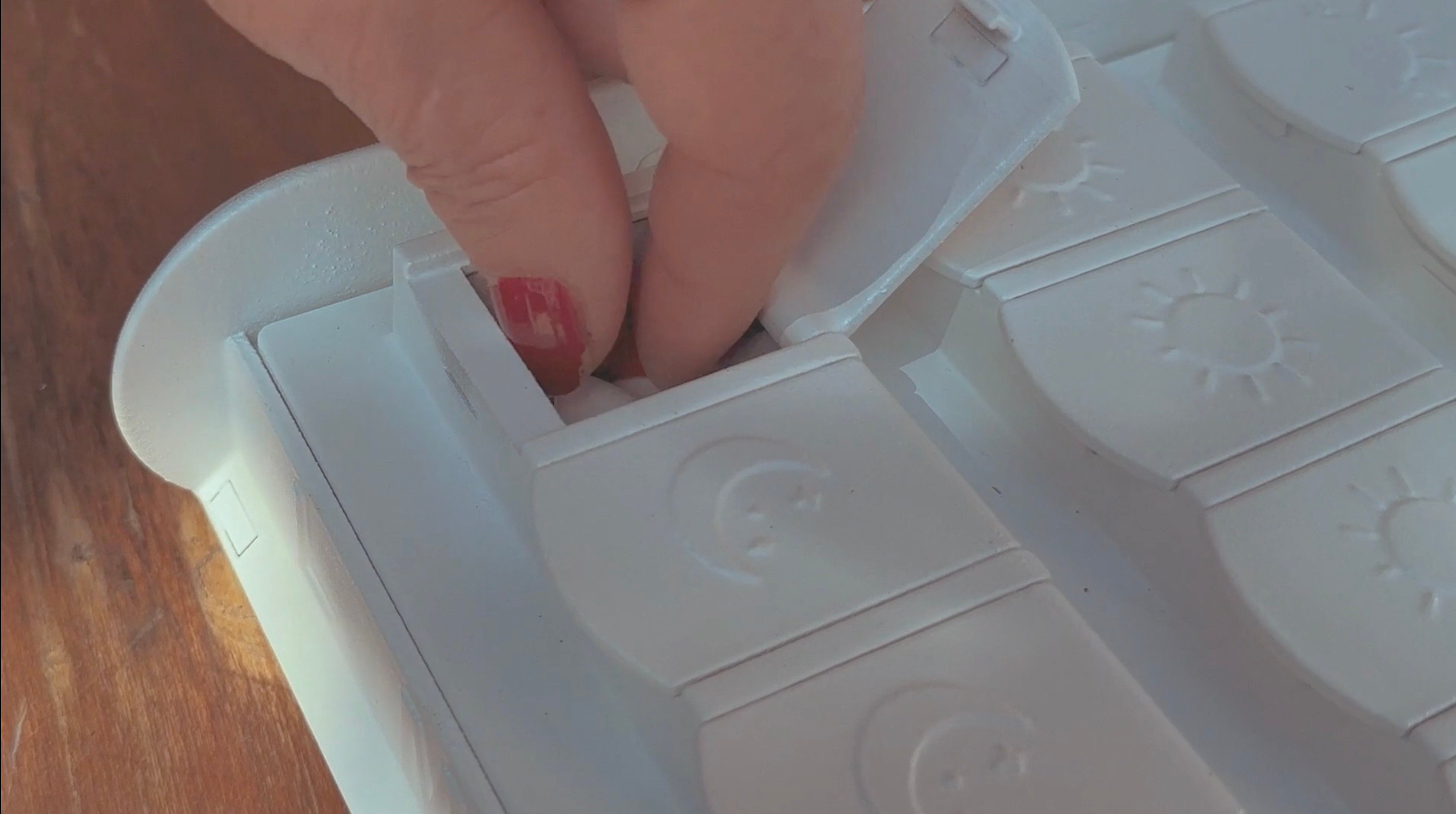  Hand opening a compartment on a white weekly pill organizer placed on a wooden surface