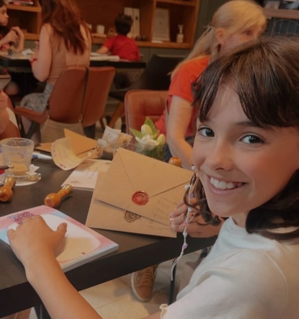 Young girl smiling at camera while holding a wax-sealed envelope at Posdata café in Buenos Aires, with letter-writing supplies on the table
