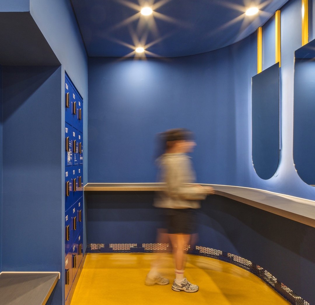 A person wearing sneakers in a brightly lit locker area with vivid blue walls, yellow flooring and lockers