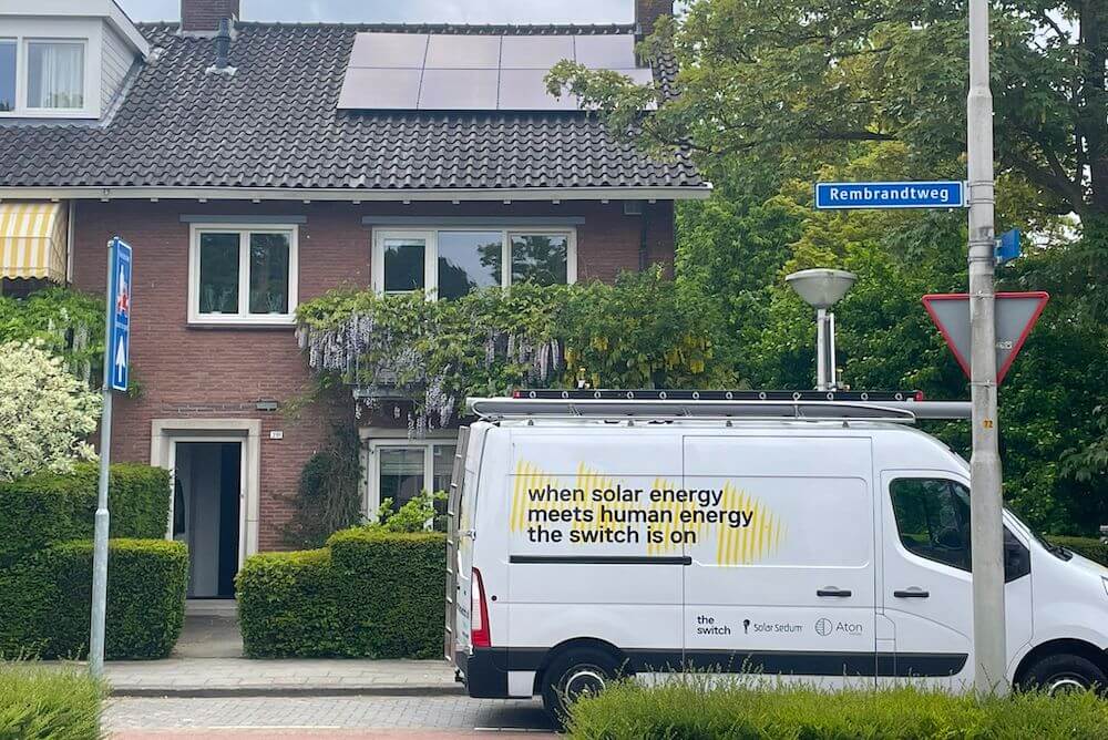 Van parked by a house with rooftop solar panels. Text on the van: 'When solar energy meets human energy the switch is on'