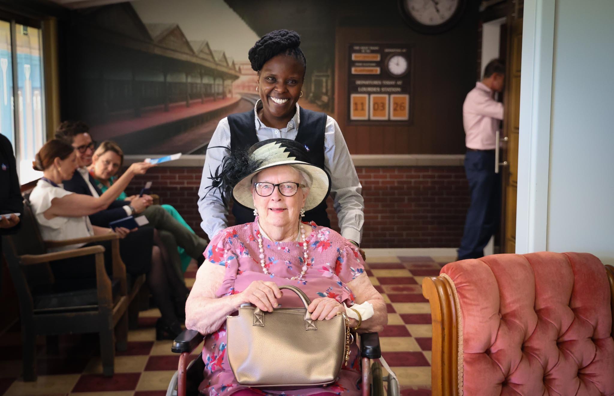 A smiling care worker pushes an elderly woman in a wheelchair through a room decorated to resemble a vintage railway station, complete with a departure board and platform mural on the wall
