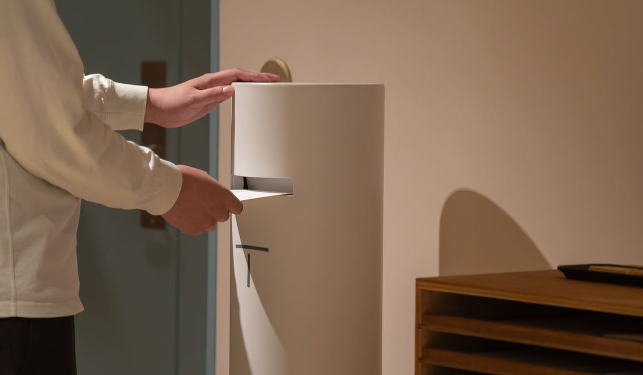 A person inserts a letter into a tall, cylindrical white postal box inside a calm, softly lit interior space