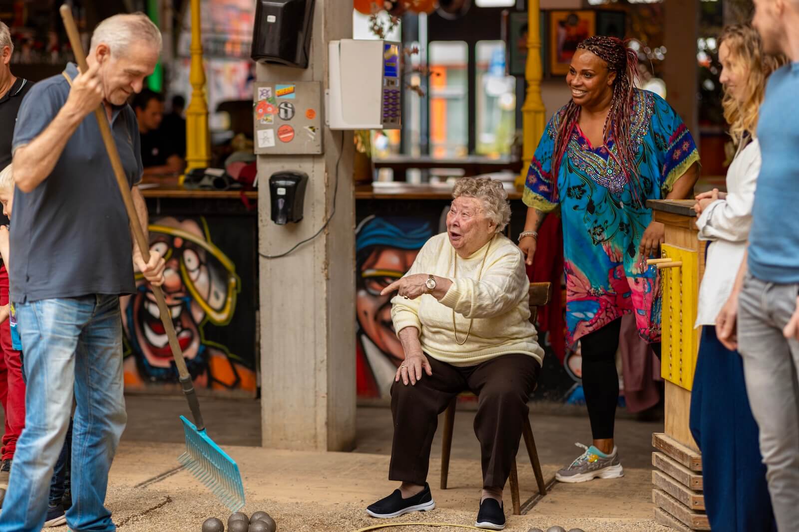 Diverse group of people playing jeu de boules in a colorful indoor venue, including an elderly woman seated in the center watching the game, with younger participants of different backgrounds standing around her in a casual, welcoming atmosphere 