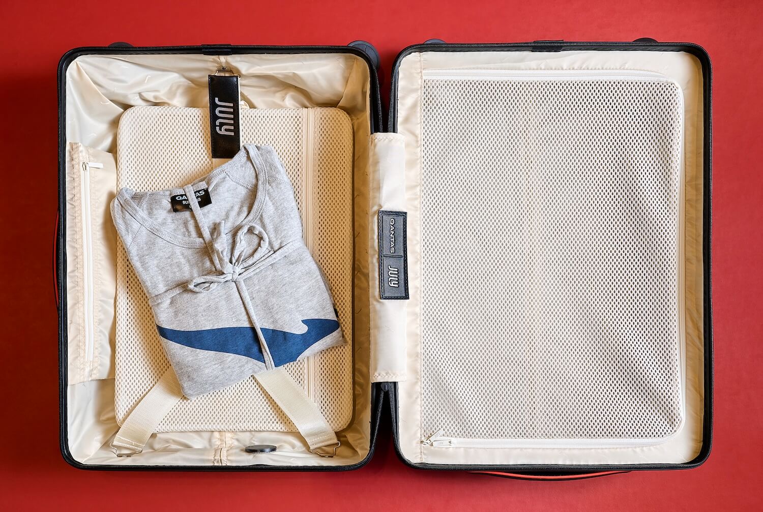 An open Qantas × July suitcase with cream interior lining and mesh compartments, displaying organized packing with a folded gray Qantas t-shirt 