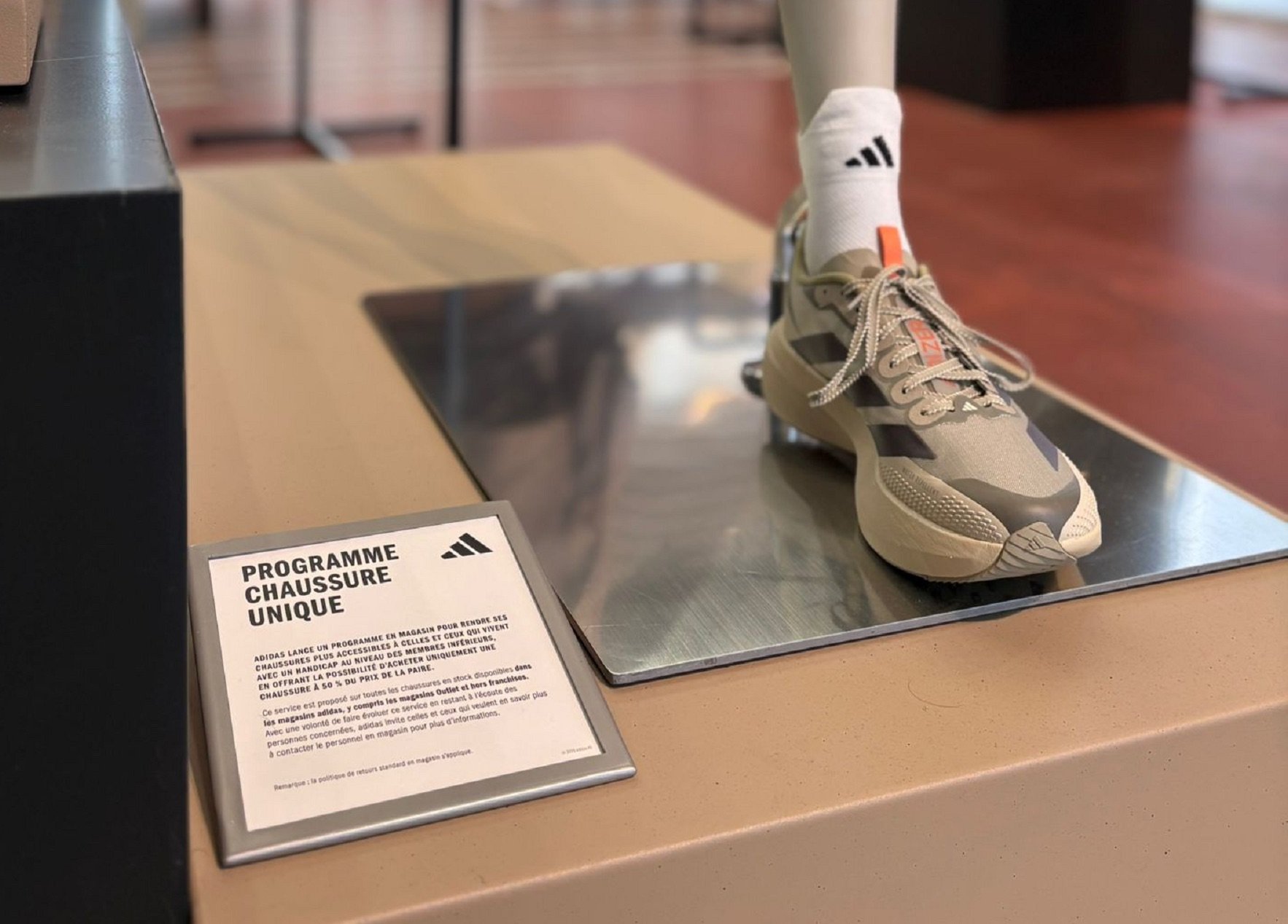 In-store display for the Adidas Single Shoe program, showing a single olive-green sneaker on a mannequin foot next to a framed information card explaining the service, in what appears to be a French Adidas store 