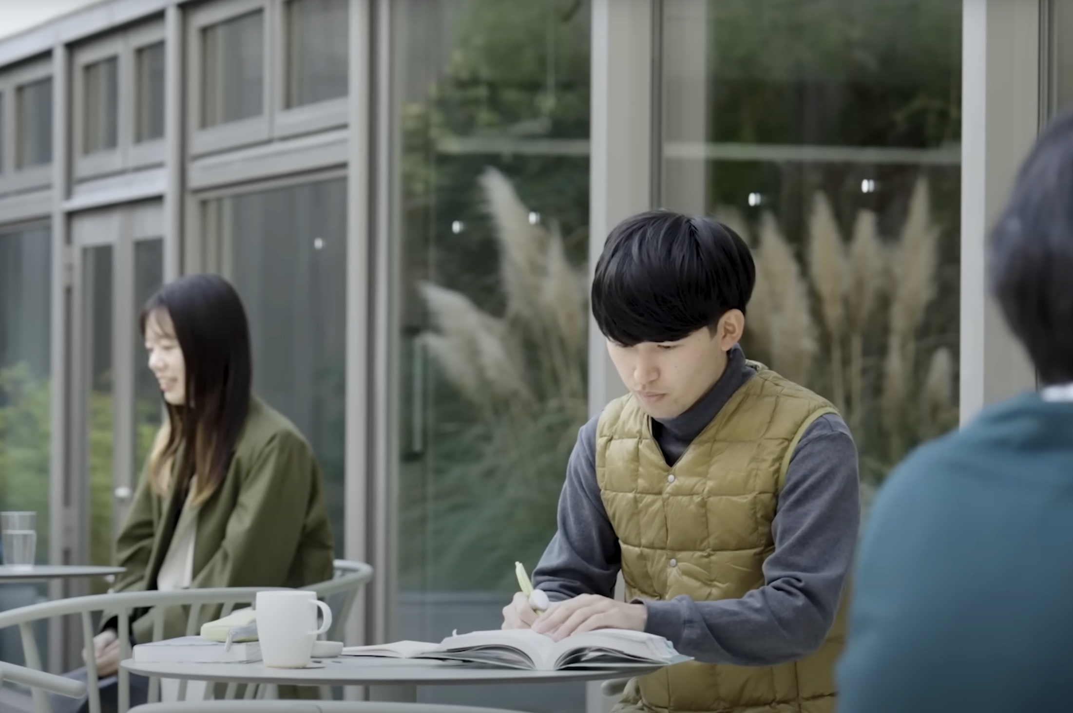 A young man sits at an outdoor café table, focused on writing in an open notebook with a pen 