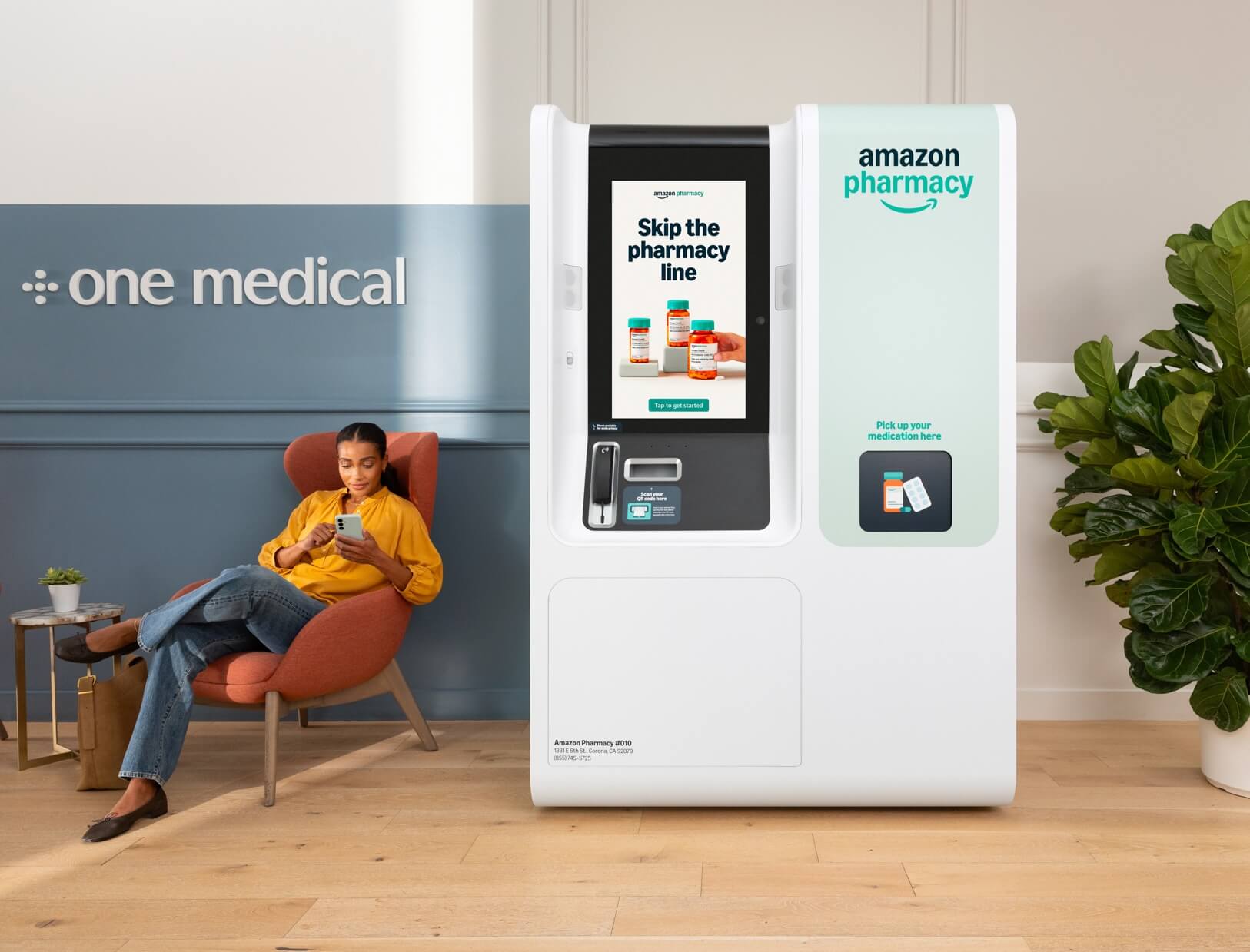 A woman sits in a clinic lounge checking her phone beside an Amazon Pharmacy kiosk at a One Medical location. The kiosk displays the message “Skip the pharmacy line” 