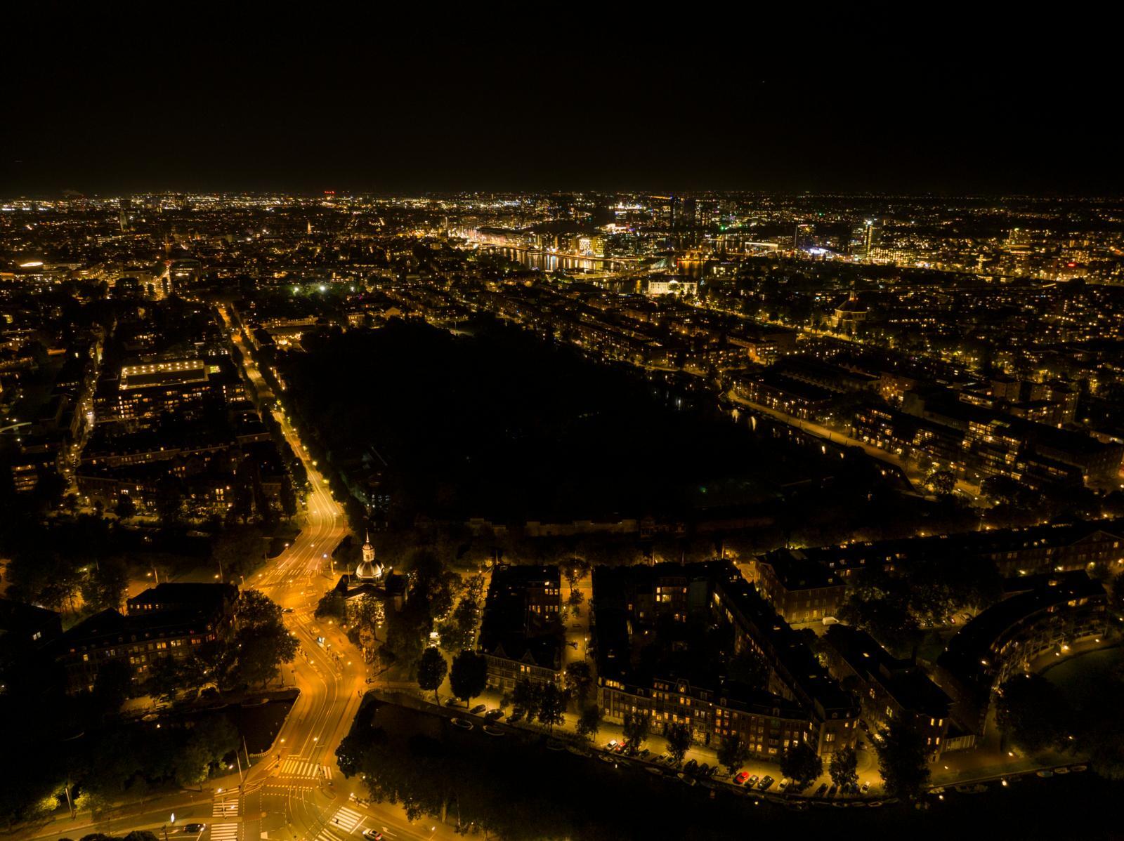 A wide, nighttime aerial view of Amsterdam lit by dense grids of yellow streetlights, with a dark, unlit zoo area forming a large patch of darkness at the center 