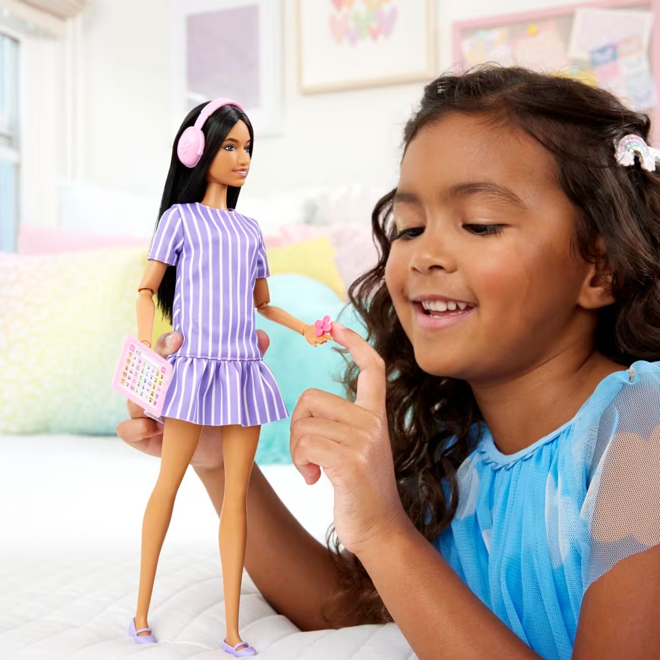 A smiling young child plays with a Barbie doll wearing pink headphones and a purple striped dress, touching the doll’s hand while sitting on a bed in a bright bedroom 