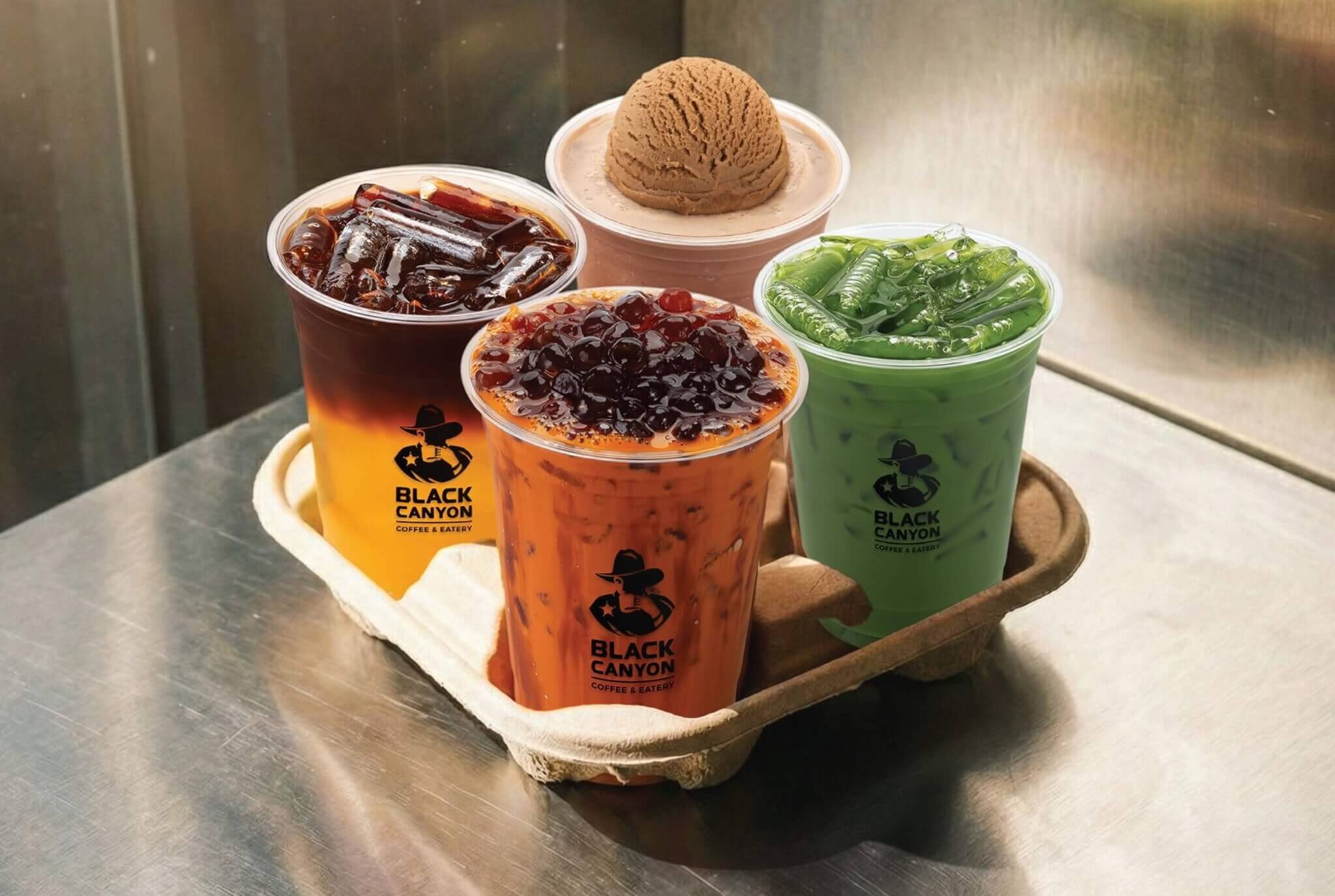 Four Black Canyon branded beverages in clear plastic cups on a wooden tray: iced tea with grass jelly, Thai milk tea with boba pearls and coffee ice cream, green tea with jelly toppings and iced coffee with chocolate grass jelly 