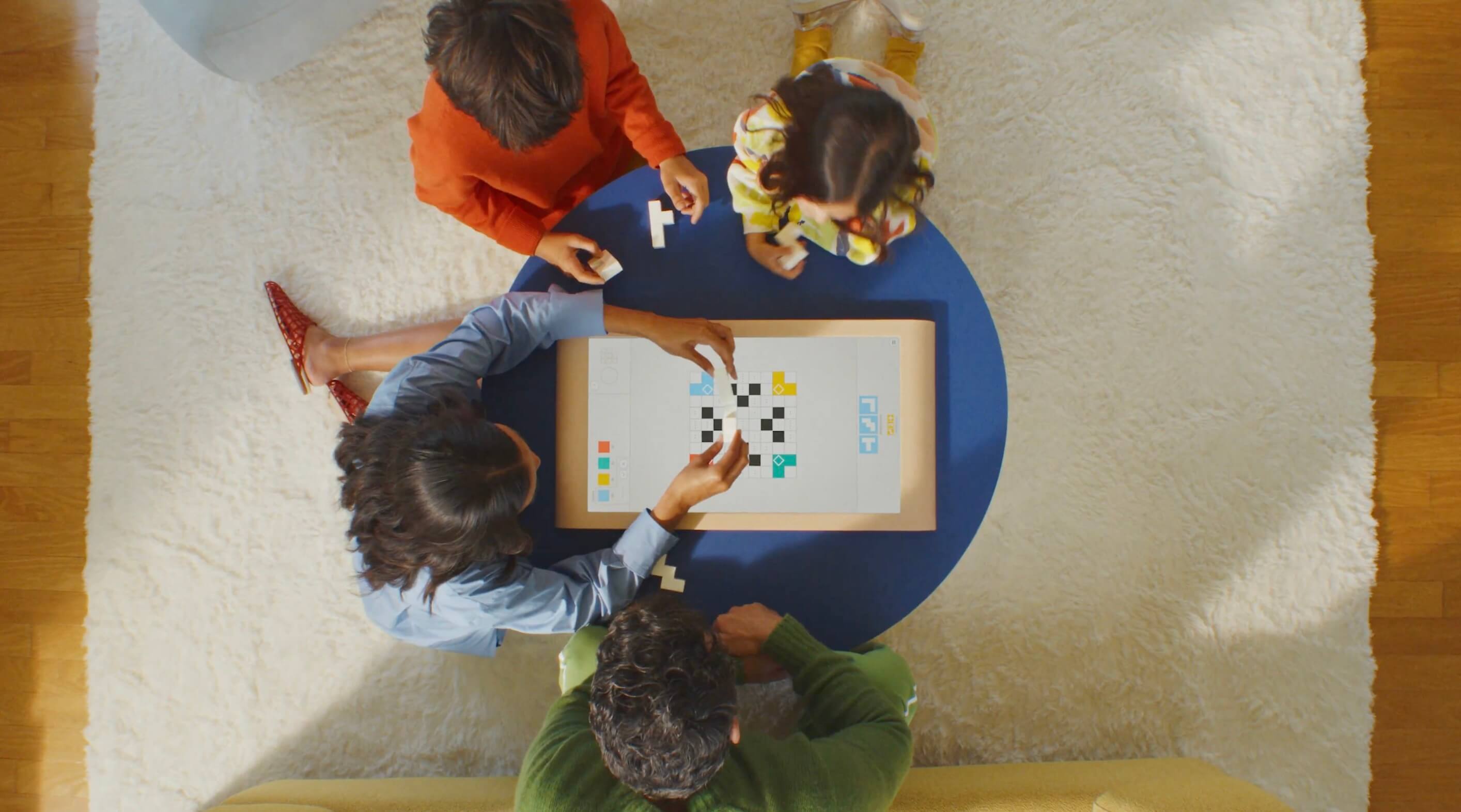 Overhead view of four people gathered around Board's gaming console. Players of different ages are seated on a cream-colored rug, reaching toward the tabletop screen with small game pieces 