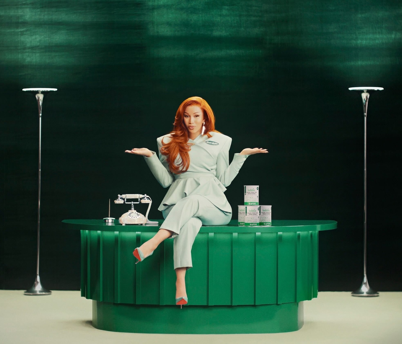 Cardi B sits on a green desk wearing a mint green power suit, arms outstretched. A vintage rotary phone and stacked Bobbie formula boxes sit on the desk beside her 