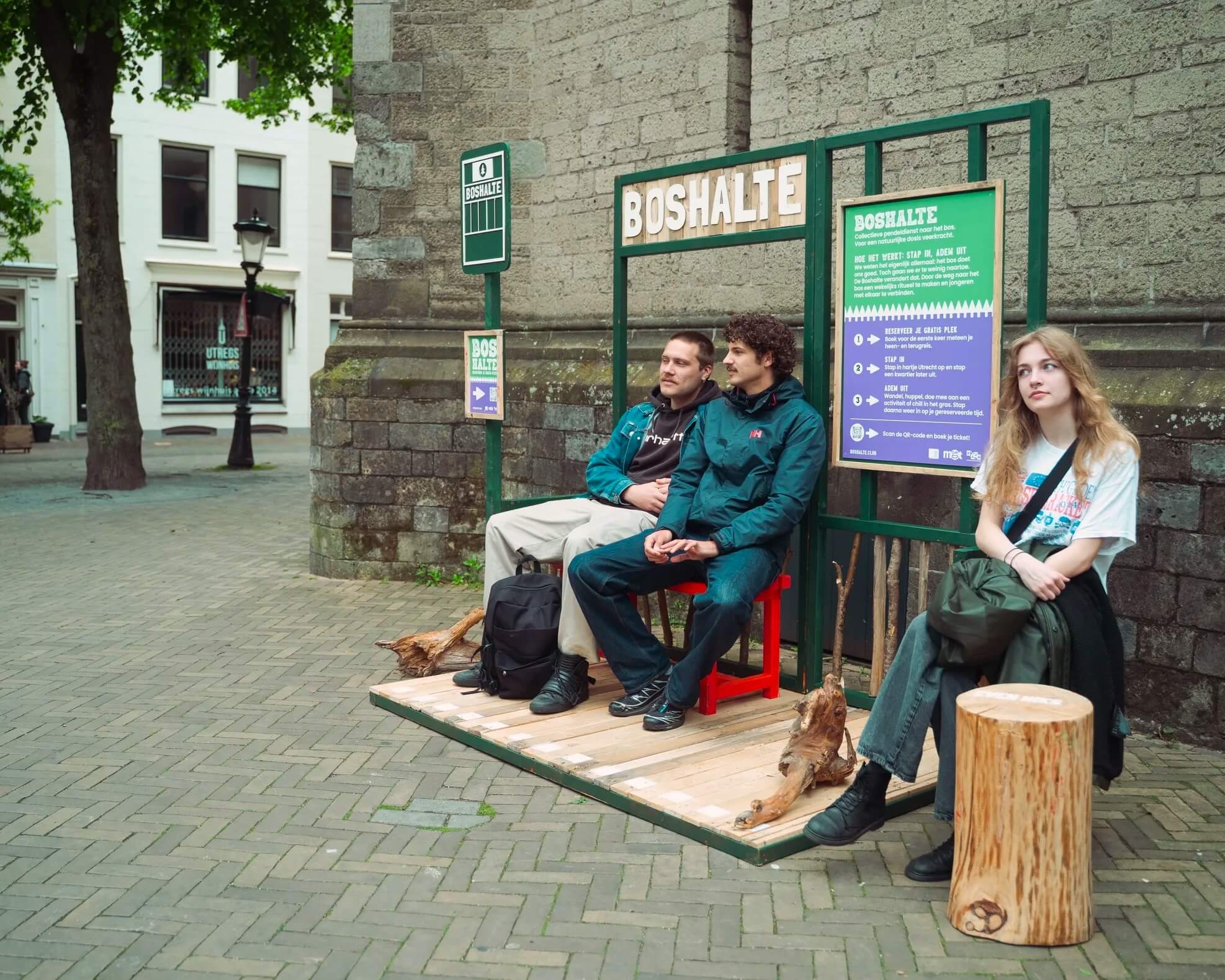 Three young adults seated at De Boshalte in central Utrecht, a small bus-stop installation with a green frame, a wooden 