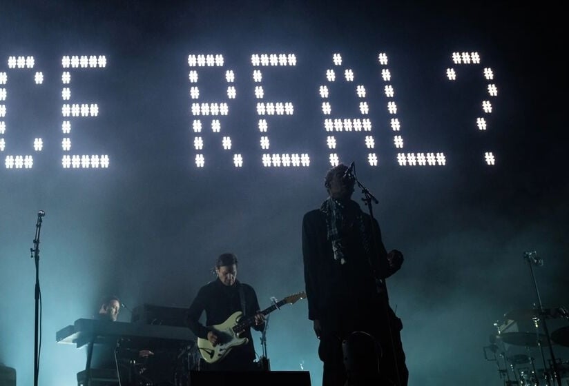 A band performs on a dimly lit stage with heavy blue haze, silhouetted against a large grid of bright white lights spelling “IS CHOICE REAL?” above them 