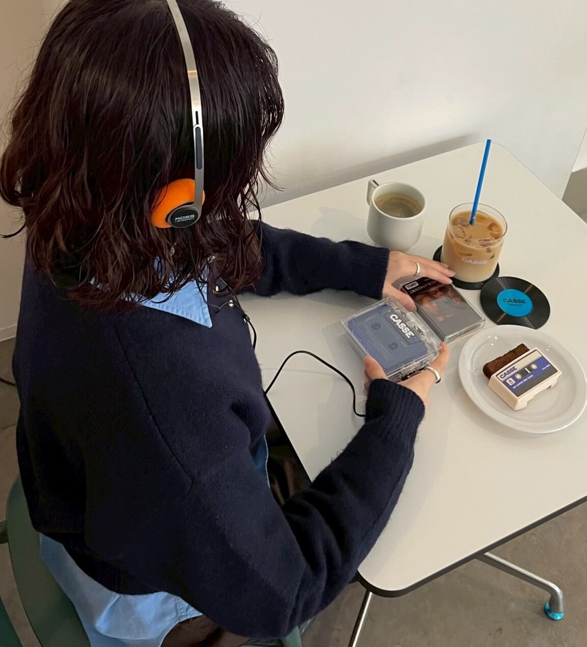 A café guest wearing headphones listens to a cassette player while holding two tapes at a white table with coffee, iced latte, and dessert in a minimalist setting 
