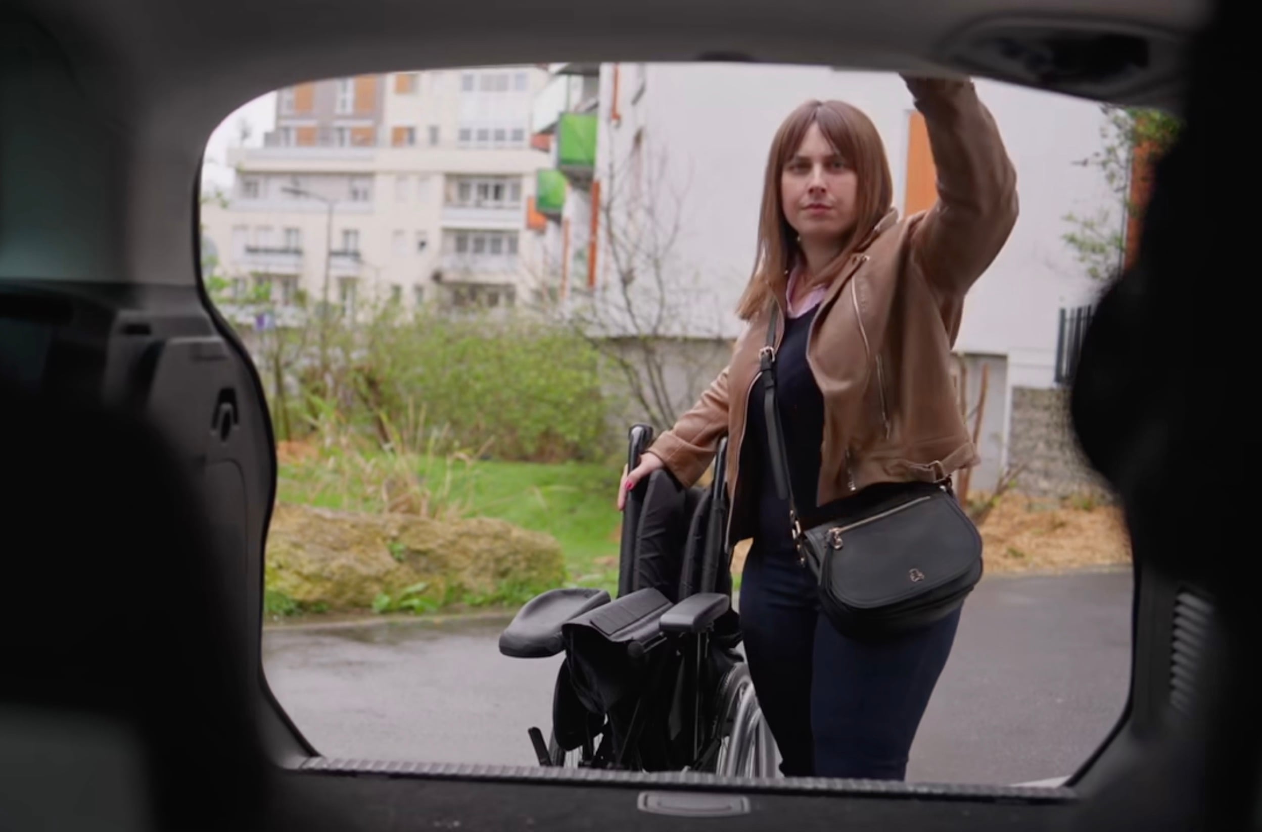 View from inside a car trunk looking out at a woman in a tan jacket lifting the trunk door while holding a folded wheelchair beside herl 