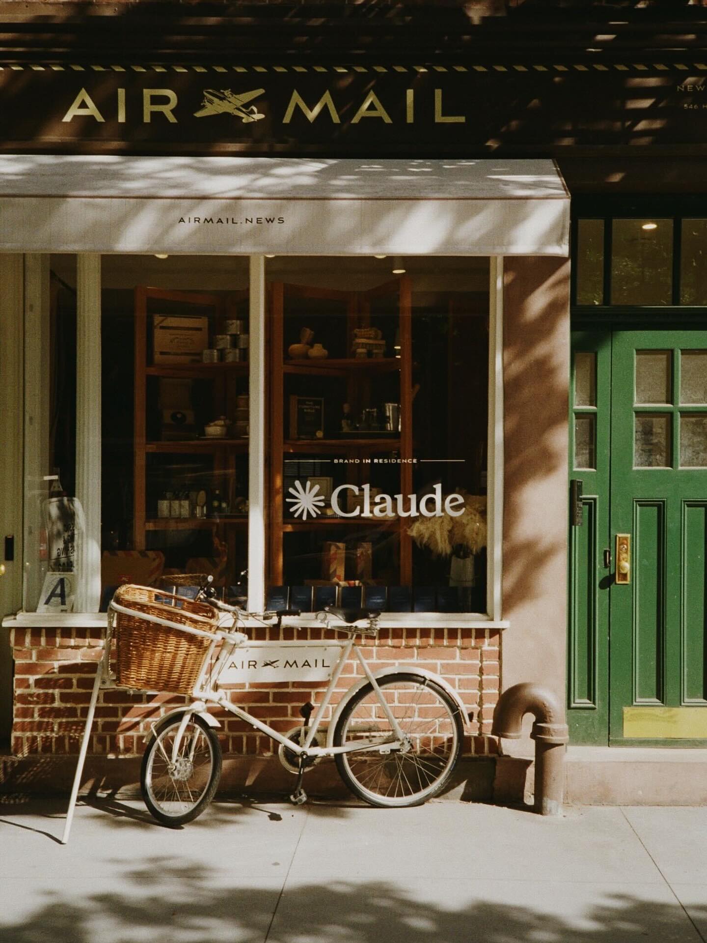 Storefront of Air Mail newsstand in NYC’s West Village featuring a window sign for Claude, Anthropic’s AI brand. A vintage-style bicycle with a wicker basket is parked out front 