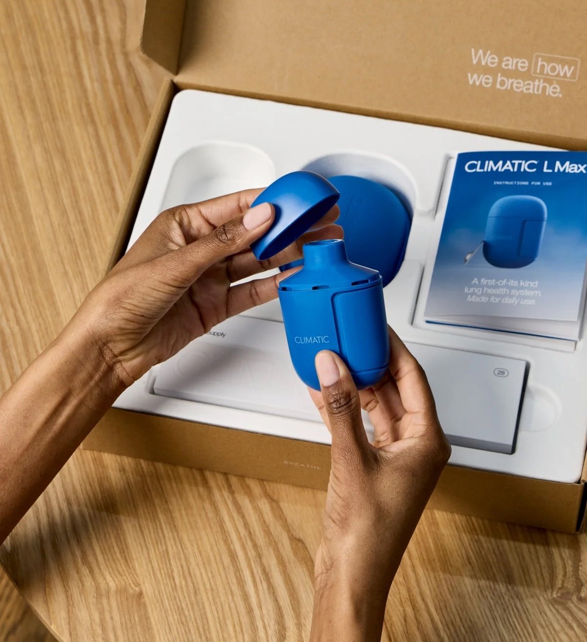 Hands holding a small blue Climatic L Max inhaler device with its cap open, above its unboxed packaging on a wooden surface 