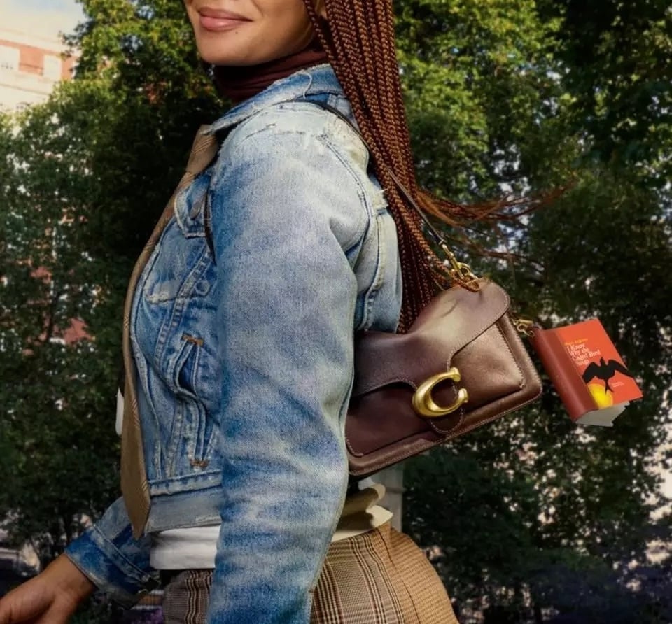 Person wearing a light blue denim jacket and a plaid skirt carries a brown leather shoulder bag with a gold clasp and a small book-shaped charm attached, walking outdoors with trees in the background 