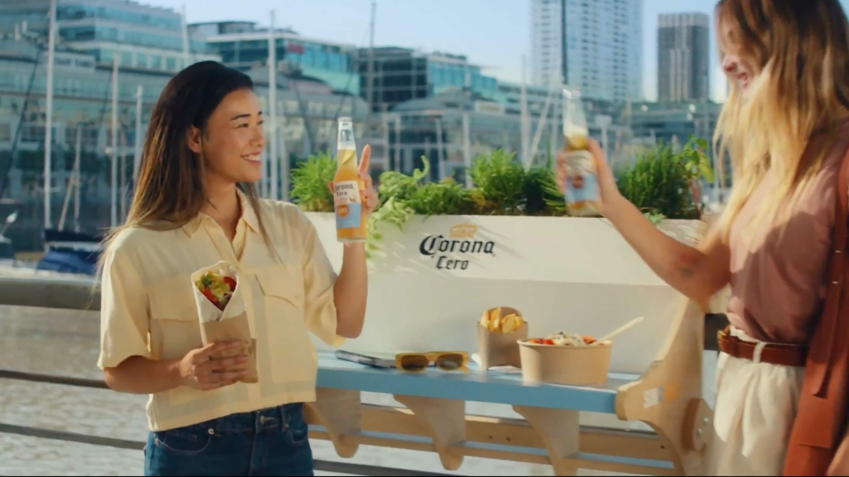 Two women sharing lunch and Corona Cero beers at a white branded table on an outdoor rooftop terrace with urban buildings in the background 