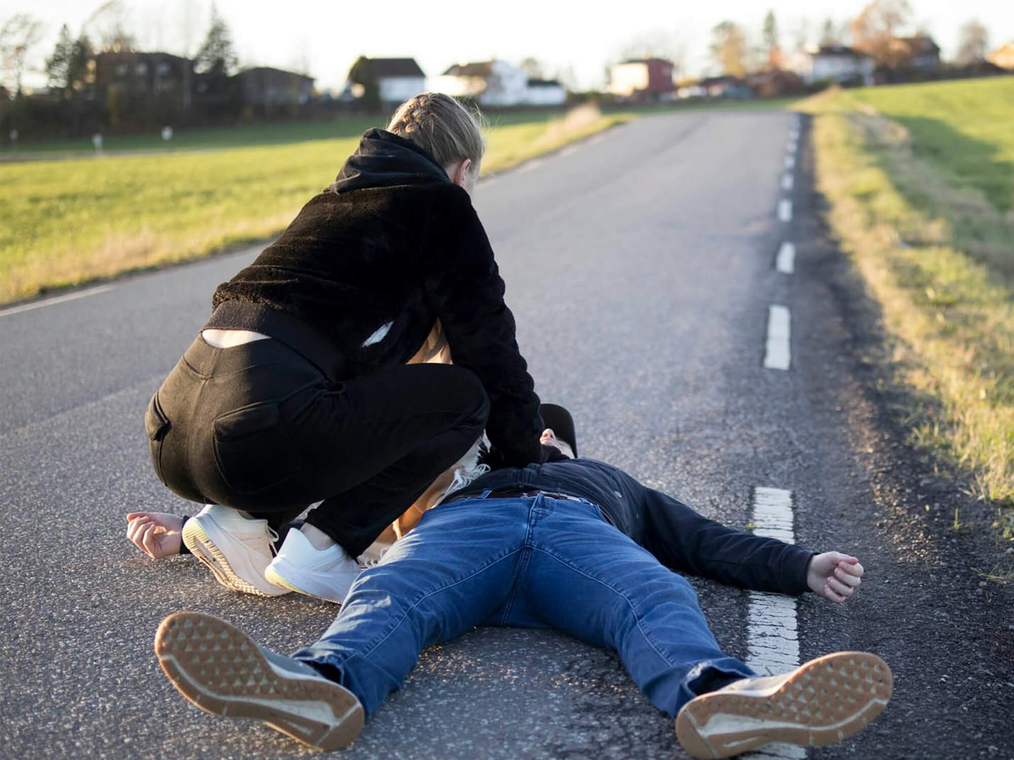 A person in a black hoodie kneels over a man lying on his back on a rural road, performing chest compressions in what appears to be a CPR scenario 