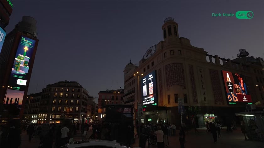 A dusk view of Plaza Callao in Madrid showing two large LED billboards displaying Plenitude advertising in dark mode, with deep-toned backgrounds visibly muted against the surrounding cityscape; a 