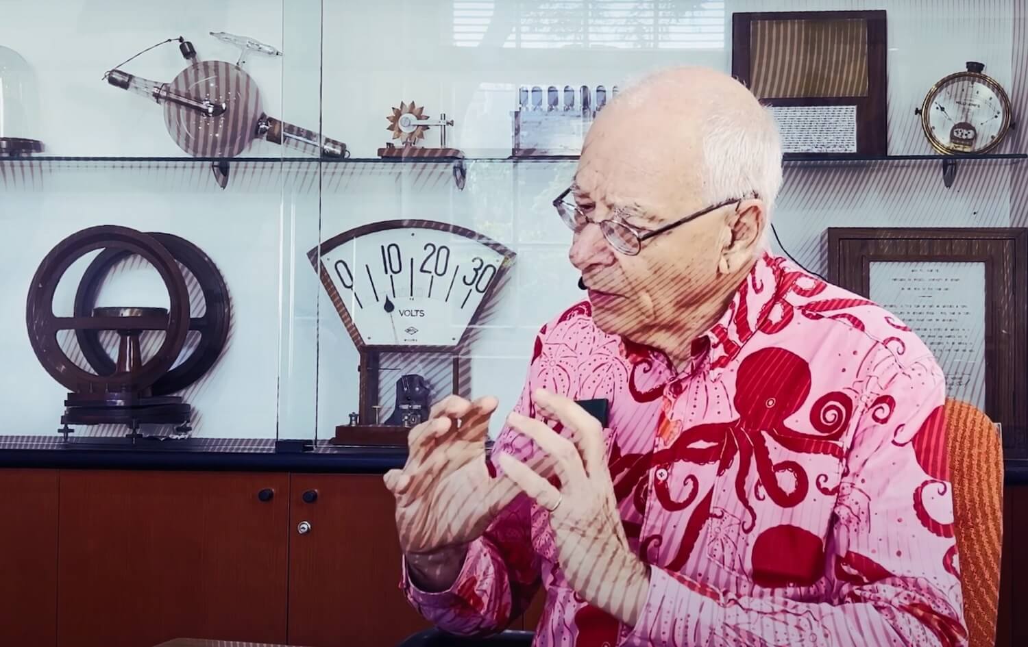 Older man with short white hair and glasses, wearing a pink shirt with a red octopus print, gesturing with his hands while speaking. He is seated in front of a glass cabinet displaying vintage scientific instruments, including a large voltmeter and various measuring devices 