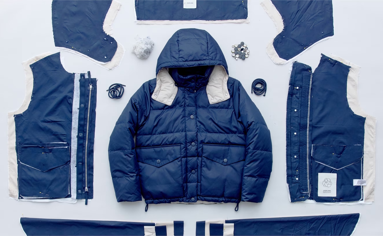 A flat-lay of a navy blue EverPuff jacket, surrounded by its disassembled components — fabric panels, zippers, buttons, cords and insulation 