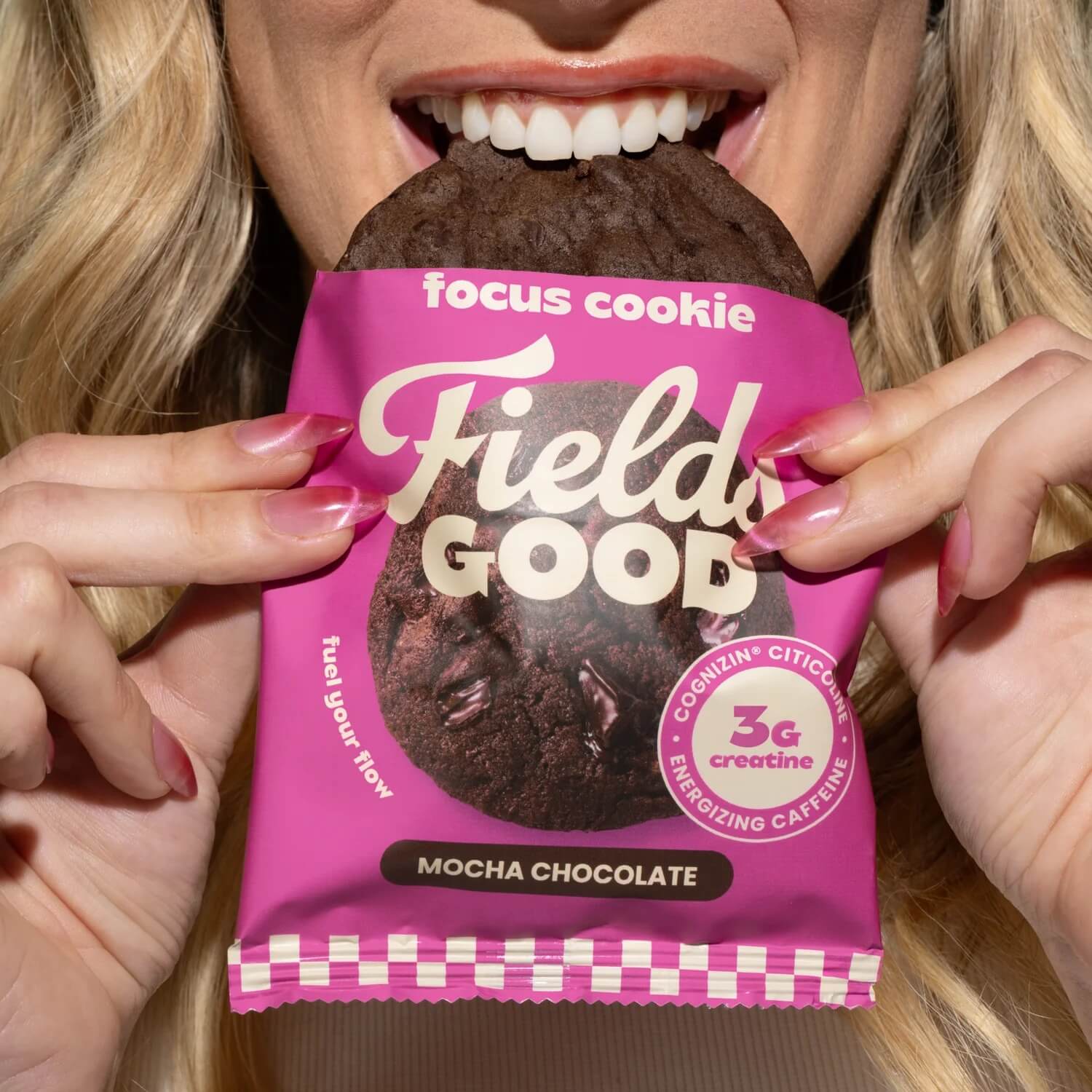 A person bites into a dark chocolate cookie held over its pink Fields Good 