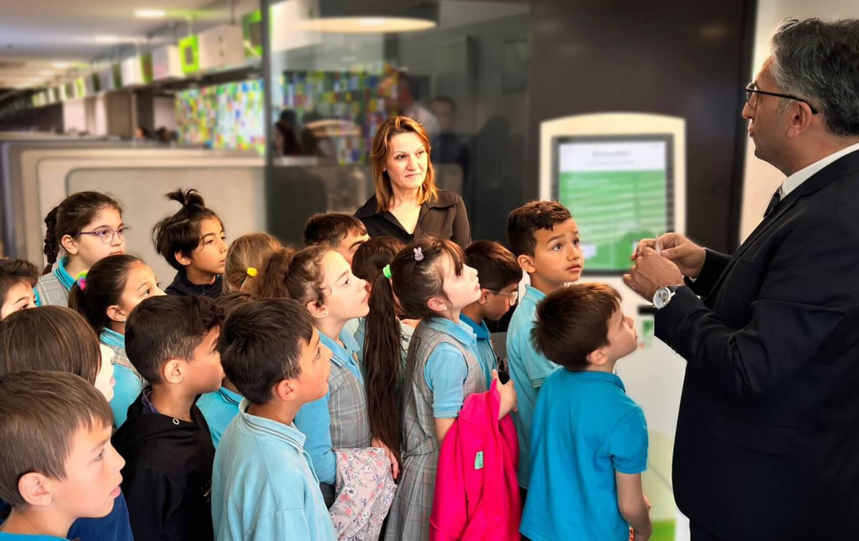 A group of young schoolchildren listen attentively to a man in a suit explaining something during a visit to a bank 