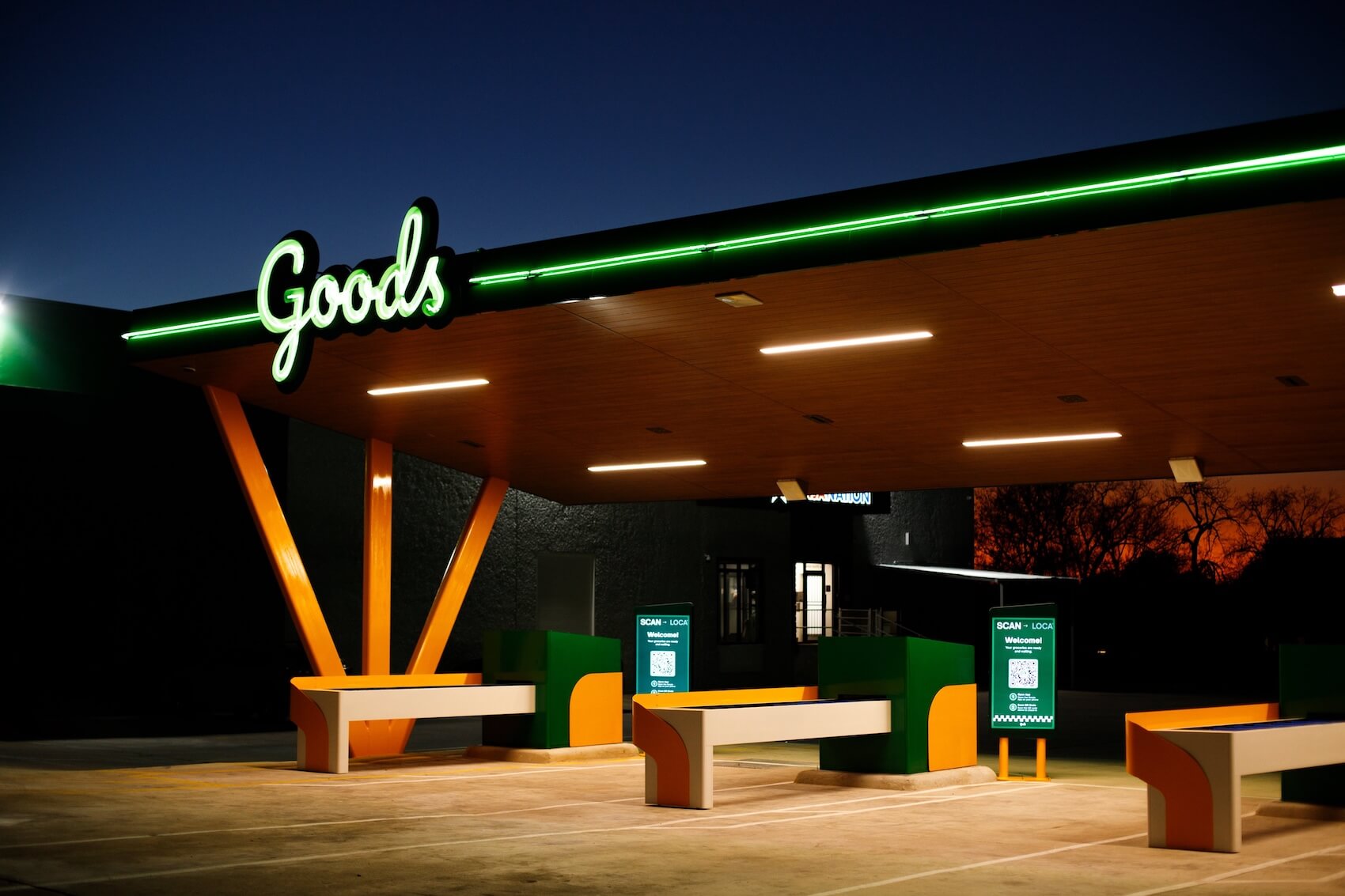 Nighttime exterior of Goods grocery store featuring a glowing green neon sign, mid-century modern canopy with orange V-shaped support columns, green and orange benches, and digital kiosk screens displaying QR codes for order pickup 