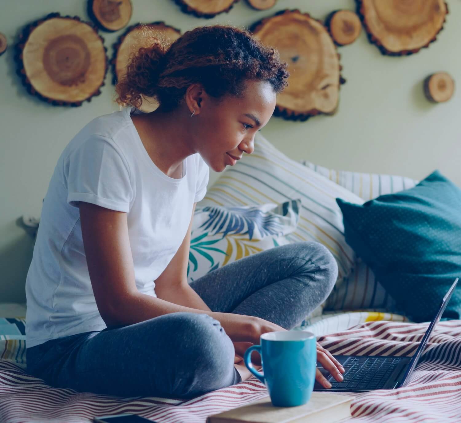 Student sitting cross-legged on a bed, focused on a laptop, with a blue mug in the foreground 