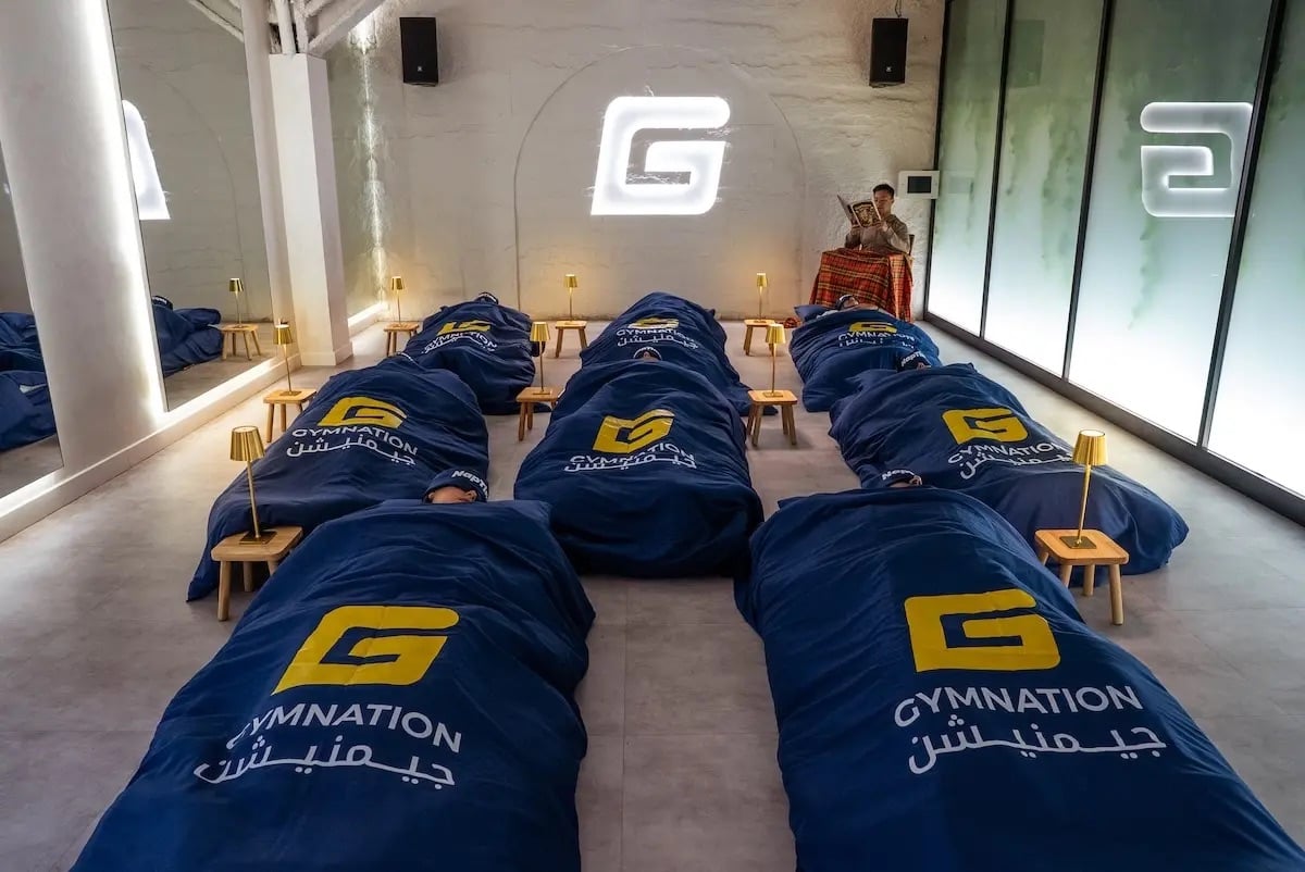 GymNation's NapTime studio with rows of people on cots with navy blue branded sleeping bags, an instructor seated in the background, and the GymNation logo projected on a textured white wall 