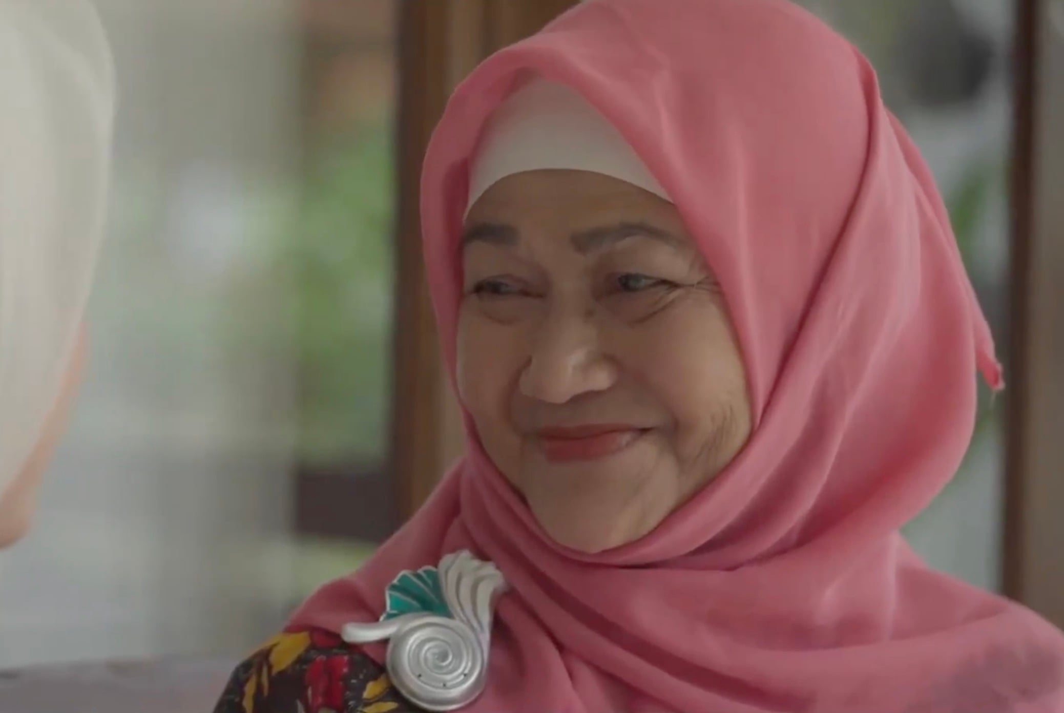 A silver and turquoise brooch shaped like a stylized snail shell, worn pinned to the pink hijab of a smiling older woman 