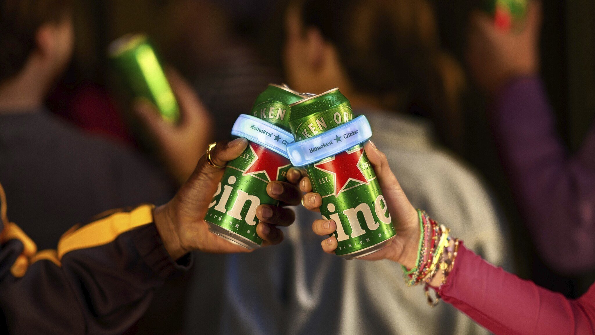 Two people clinking Heineken cans fitted with light-up Clinker bands at a festival, with a crowd visible in the background 