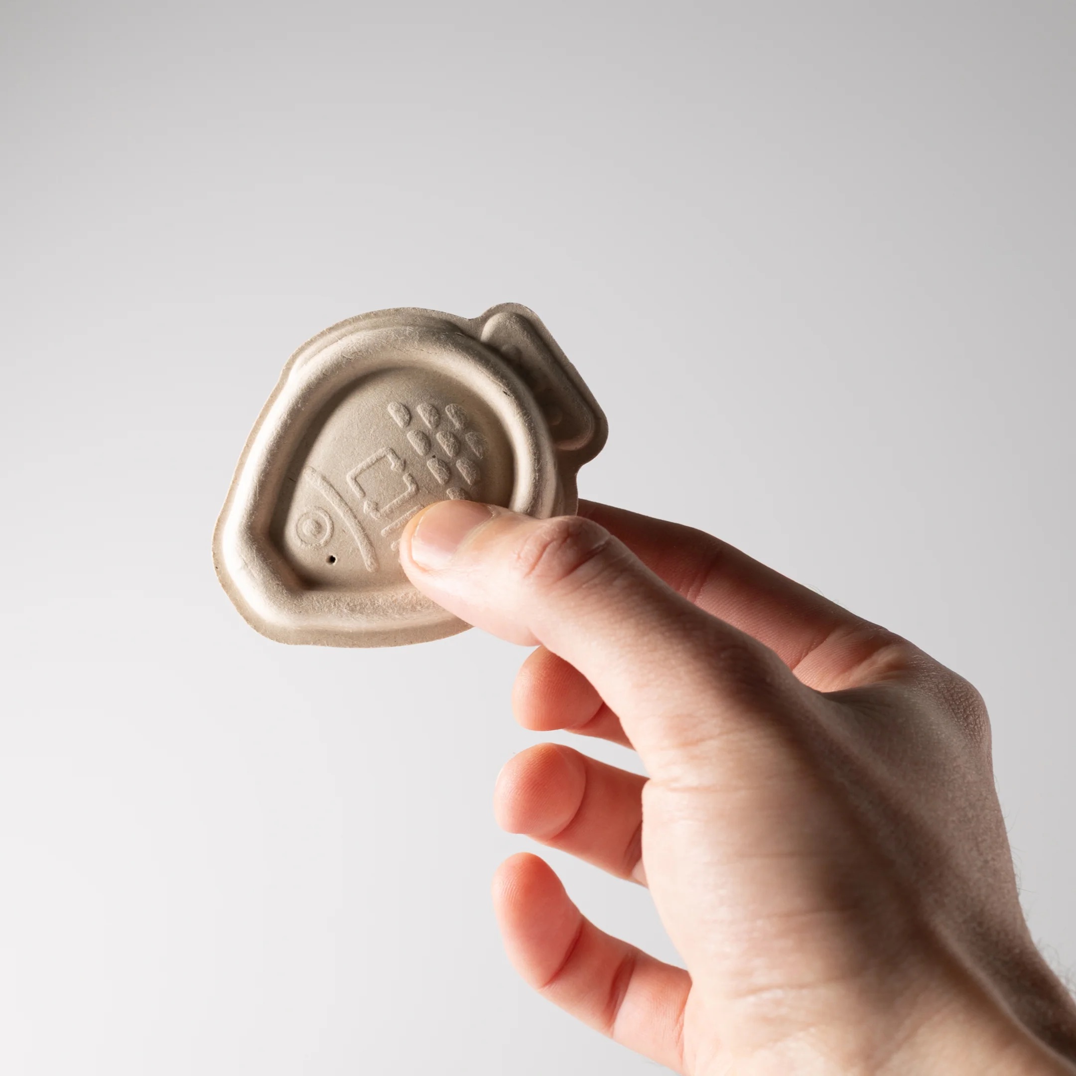 A hand holds a beige, fish-shaped soy sauce dropper made from compostable bagasse plant pulp 