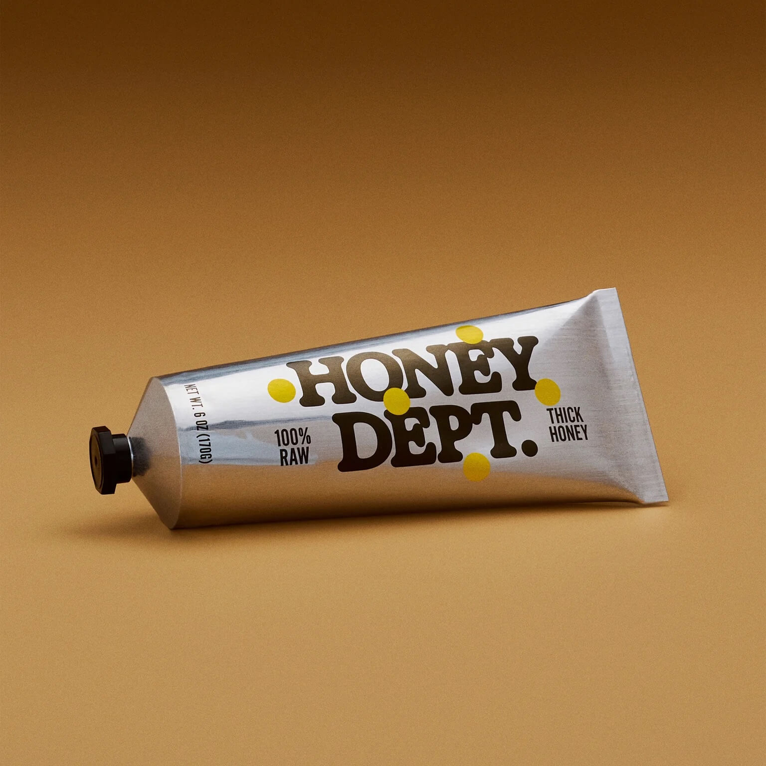 Silver aluminum squeeze tube labeled “Honey Dept. Thick Honey” with yellow dot accents and black cap, photographed on a warm tan background 