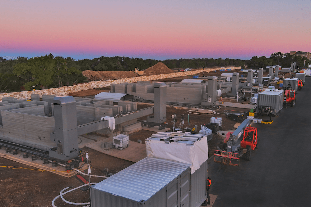A row of five huge robots 3D printing homes, with a pink sky in the background