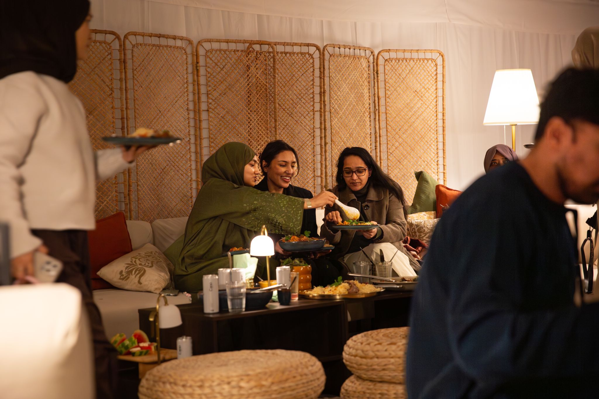 Group of people sharing food from communal dishes in warmly lit living room setting with woven room divider and floor cushions 