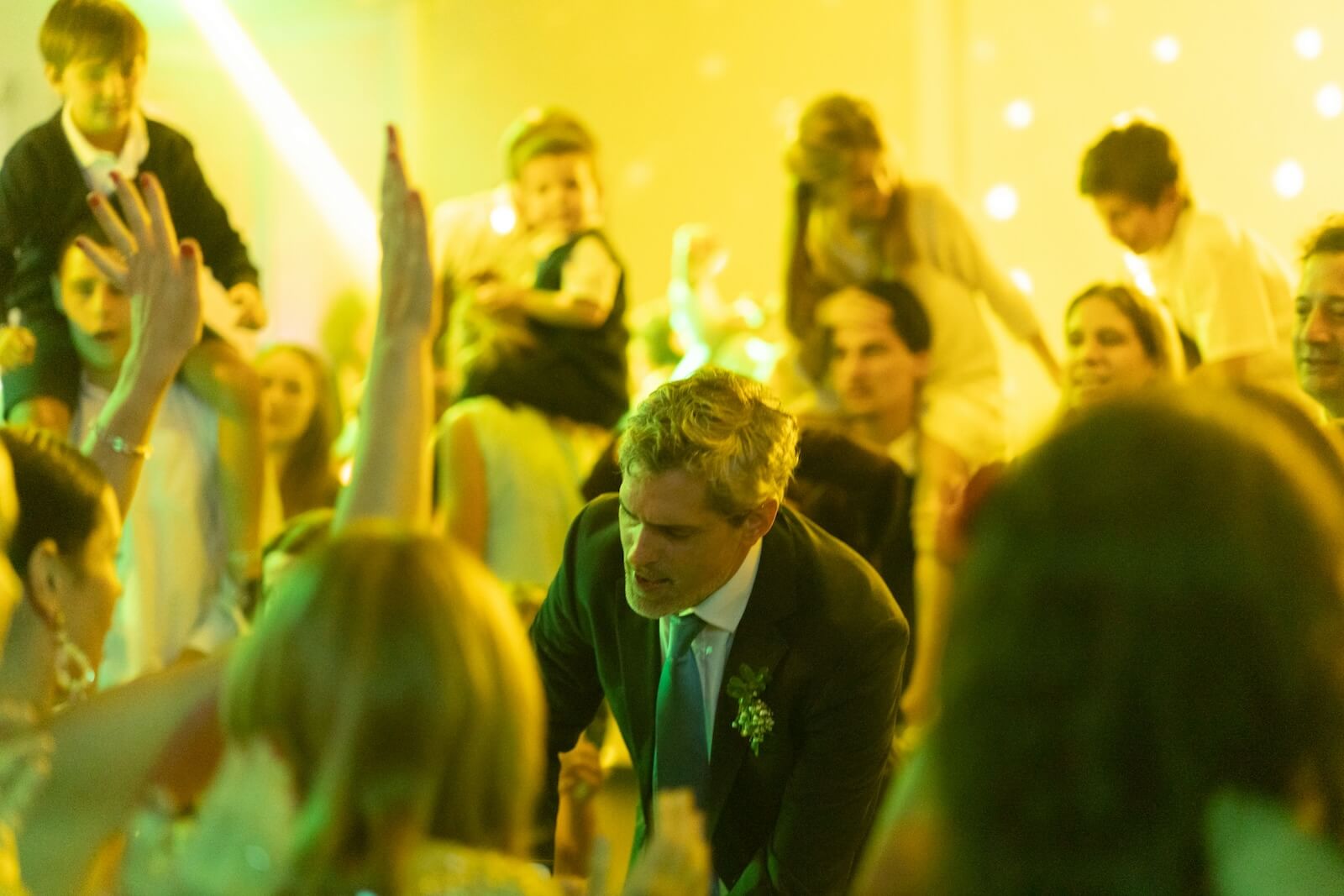 A lively celebration scene with a crowd of people dancing and enjoying themselves under green and yellow lighting 