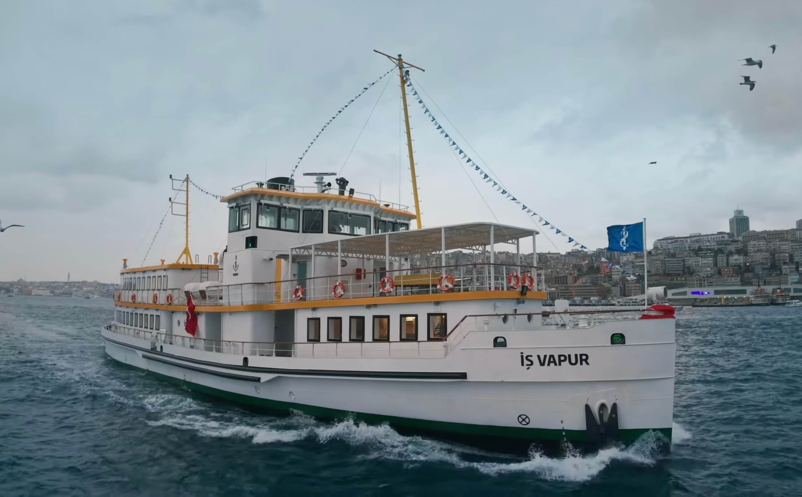 White-and-yellow passenger ferry named “İş Vapur” sailing on choppy water, decorated with blue pennant flags, with seagulls overhead and Istanbul’s shoreline in the background 