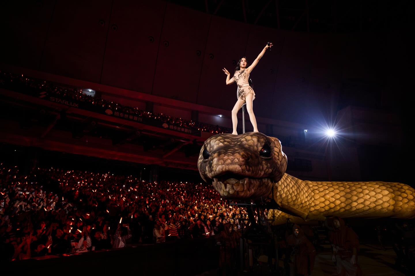 A pop singer stands atop a massive mechanical serpent inside a packed indoor arena, illuminated by stage lights as thousands of fans hold glowing light sticks below 