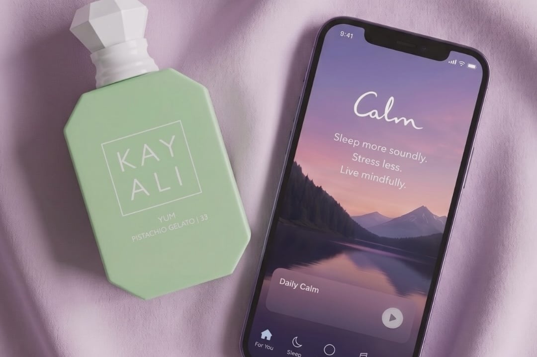 A pastel green Kayali Yum Pistachio Gelato perfume bottle lies on a lilac fabric next to a smartphone displaying the Calm app's home screen 