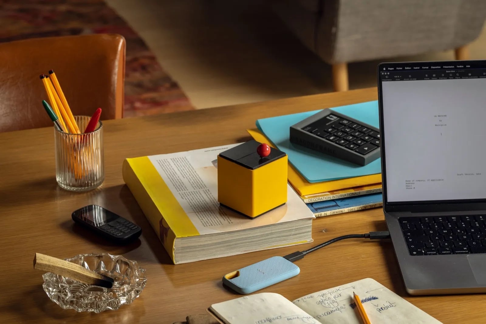 A yellow cube with a black lid and a red button sits on a stack of magazines on a wooden desk, next to an open laptop, a small black dumbphone, a glass of pencils, an external drive, and a crystal ashtray holding a palo santo stick 