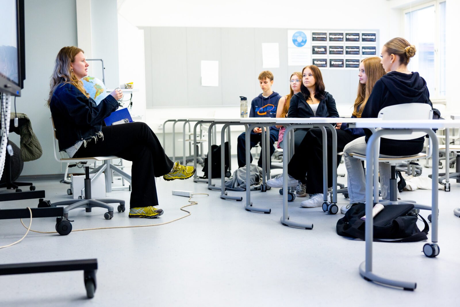 A young woman sits in a classroom, speaking to a group of high school students 